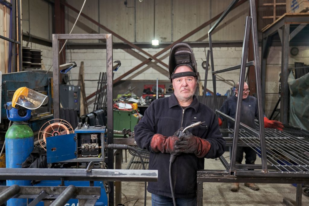 Erfahrener Metallbauer mit Schweißhelm in der Werkstatt neben Stahlrahmen