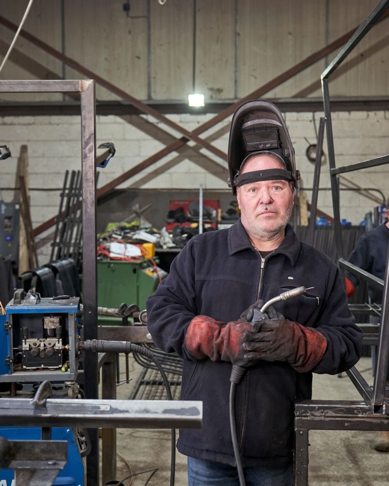 Erfahrener Metallbauer mit Schweißhelm in der Werkstatt neben Stahlrahmen